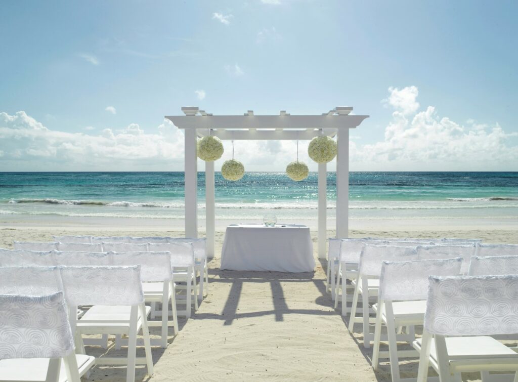beach wedding ceremony set up with a white pergola with hanging flowers and white chairs