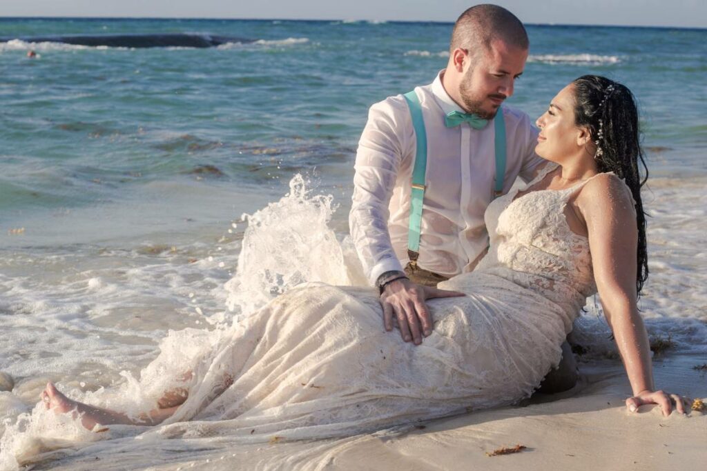 Just married couple looking at each other sitting on a beach getting hit by the waves