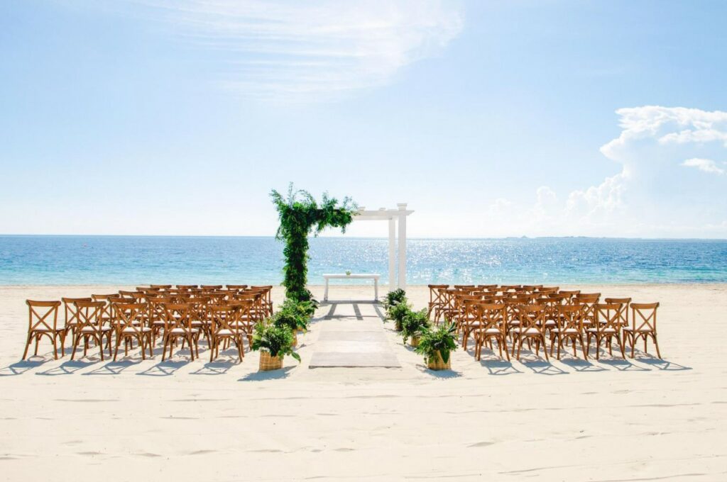Beach wedding set up with a white pergola with greenery and wooden chairs